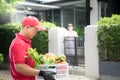 Asian delivery man in red uniform delivering  groceries box of food, fruit, vegetable and drink to woman recipient at home Royalty Free Stock Photo