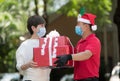 Asian delivery man wearing face mask and gloves in red uniform and Christmas hat delivering present and gift boxes to woman recipi Royalty Free Stock Photo