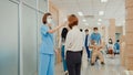 Asian doctor walk through hospital hallway with helper staff push patient wheelchair, operator check fever by digital thermometer