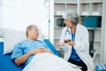 Asian doctor in white suit take notes while discussing and Asian elderly, man patient who lying on bed with receiving saline Royalty Free Stock Photo