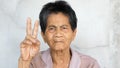 Asian elderly native woman showing two fingers gesture for encouragement with smile, looking to camera. Senior female have relaxin