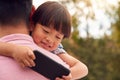 Asian Father Cuddling Son In Garden As Boy Looks Over His Shoulder At Mobile Phone