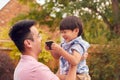 Asian Father Cuddling Son In Garden As Boy Plays With Mobile Phone