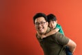 Asian father giving son ride on the back with a red background. Portrait of a happy father giving son piggyback a ride on his Royalty Free Stock Photo