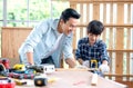 Asian father look carefully son use saw with timber in their workplace of carpentering with happy emotion. Asian family concept to