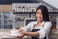 Asian female barista hold a cup of coffee serving to her customer with smile surrounded with bar counter background. Royalty Free Stock Photo