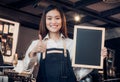 Asian female barista wear jean apron thumbs up at blank blackboard coffee menu at counter bar with smile face,cafe service