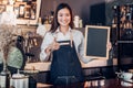 Asian female barista wear jean apron thumbs up at blank blackboard coffee menu at counter bar with smile face,cafe service