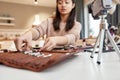 Asian female blogger showing cosmetic products and makeup brushes while recording a tutorial video for her beauty blog Royalty Free Stock Photo