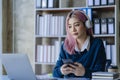 Asian female college student wearing headphones studying online using mobile smartphone app contacting laptop