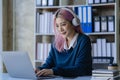 Asian female college student wearing headphones studying online using mobile smartphone app contacting laptop