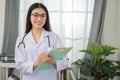 Asian female doctor with stethoscope holding clipboard standing looking at camera and smiling happily in her office at clinic Royalty Free Stock Photo