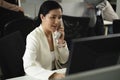 Asian female employee with white suit sit in black chair busy with hotline front of monitor and many queue of co-worker behind in Royalty Free Stock Photo