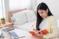 Asian female freelancer reading and jot down note on book while working with laptop on table in bedroom at home.Work at home