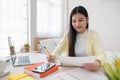 Asian female freelancer reading report on laptop and jot down note on table in bedroom at home.Work at home concept.work from home