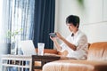 Asian female freelancer sitting on couch taking photo with smartphone & working on laptop at home Royalty Free Stock Photo