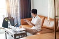 Asian female freelancer sitting on couch taking photo with smartphone & working on laptop at home