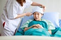 Asian female long hair doctor in white lab coat stand put cool gel pad on young sick ill girl patient wears green hospital uniform Royalty Free Stock Photo