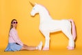 asian female model in sunglasses sitting on floor and decorative unicorn on yellow
