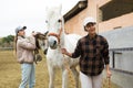 Asian female stable owner holding racehorse by bridle outdoor while girl putting saddle Royalty Free Stock Photo