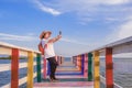 Asian female tourist using smartphone to taking selfie on rainbow wooden bridge at sea viewpoint against blue sky Royalty Free Stock Photo