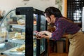 Asian female worker wearing apron holding bakery tongs wooden tray in hand, walk over refrigerated cake cabinet, open cabinet use