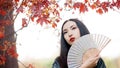 Beautiful young Asian woman with fan on background of red maple Royalty Free Stock Photo