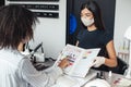 Asian girl in protective mask looks at african american client, chooses color in nail studio interior