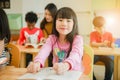 Asian girl reading a book smiling at the camera. Row of multiethnic elementary students reading book in classroom at school.