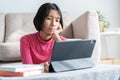 Asian girl is sitting thinking and reviewing lessons at tablet with hand on face in the living room at home morning and sunlight.