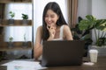 Asian girl typing data to her computer notebook for prepare her report on the wooden deak with the green plant on the table