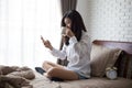 Asian girl using smartphone send Message , woman holds cup of coffee in hands on her bed In white bedroom by the window Royalty Free Stock Photo