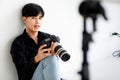 Asian handsome male model wearing casual black shirt with jeans, holding a camera, sitting on white background in a studio and Royalty Free Stock Photo