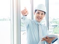 Asian happy architect or engineer man wearing white safety helmet pointing finger up, inspecting construction on huge window glass Royalty Free Stock Photo