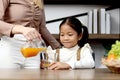 Asian happy family spending time together, mother pouring fresh orange juice from jug for her little cute daughter sitting in Royalty Free Stock Photo