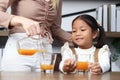 Asian happy family spending time together, mother pouring fresh orange juice from jug for her little cute daughter sitting in Royalty Free Stock Photo