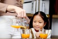Asian happy family spending time together, mother pouring fresh orange juice from jug for her little cute daughter sitting in Royalty Free Stock Photo