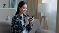 Asian happy smiling girl woman sitting home sofa looking at mobile phone chatting browsing using wifi web service Royalty Free Stock Photo