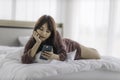 Asian happy woman sitting on bed is enjoying listening to music and watching entertainment from her smartphone with earphones Royalty Free Stock Photo