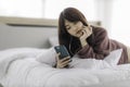 Asian happy woman sitting on bed is enjoying listening to music and watching entertainment from her smartphone with earphones Royalty Free Stock Photo