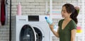 Asian housewife looking at fabric softener bottle in front of the washing machine. Working atmosphere in the laundry room