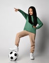 Asian kid girl in green sportswear and white sneakers stands with soccer football ball under her foot, pointing finger Royalty Free Stock Photo