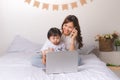 Asian lady in classic suit is talking on the mobile phone and working on laptop at home with her baby girl. Royalty Free Stock Photo