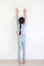 Asian little child girl raise hand up and climbing posture at white wall of room. Back view