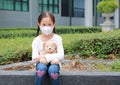 Asian little child girl wearing mask to protect corona virus and air pollution dust with hugging teddy bear doll sitting outdoor. Royalty Free Stock Photo