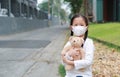 Asian little child girl wearing mask to protect corona virus and air pollution dust with hugging teddy bear doll sitting outdoor. Royalty Free Stock Photo