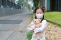 Asian little child girl wearing mask to protect corona virus and air pollution dust with hugging teddy bear doll sitting outdoor. Royalty Free Stock Photo