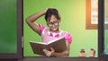 Asian little dark skinned girl in eyeglasses scratching her head in doubtful and suspect gesture while reading textbook in glass