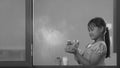 Asian little dark-skinned girl using spray bottle spraying water into her cactus plant inside of glass window frame at home in Royalty Free Stock Photo