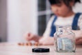 Asian Little Asian Girl Child Is Putting Thai Bath Coins And Banknote Into Piggy Bank For Saving Money On Wooden Table. Child